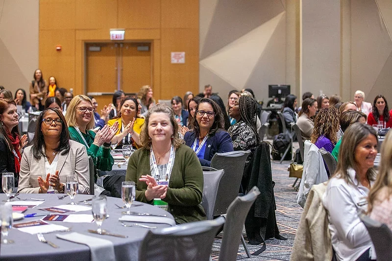 Audience of women clapping and engaging during a conference event at a large venue, with tables set for dining and a backdrop of attendees in a professional setting.